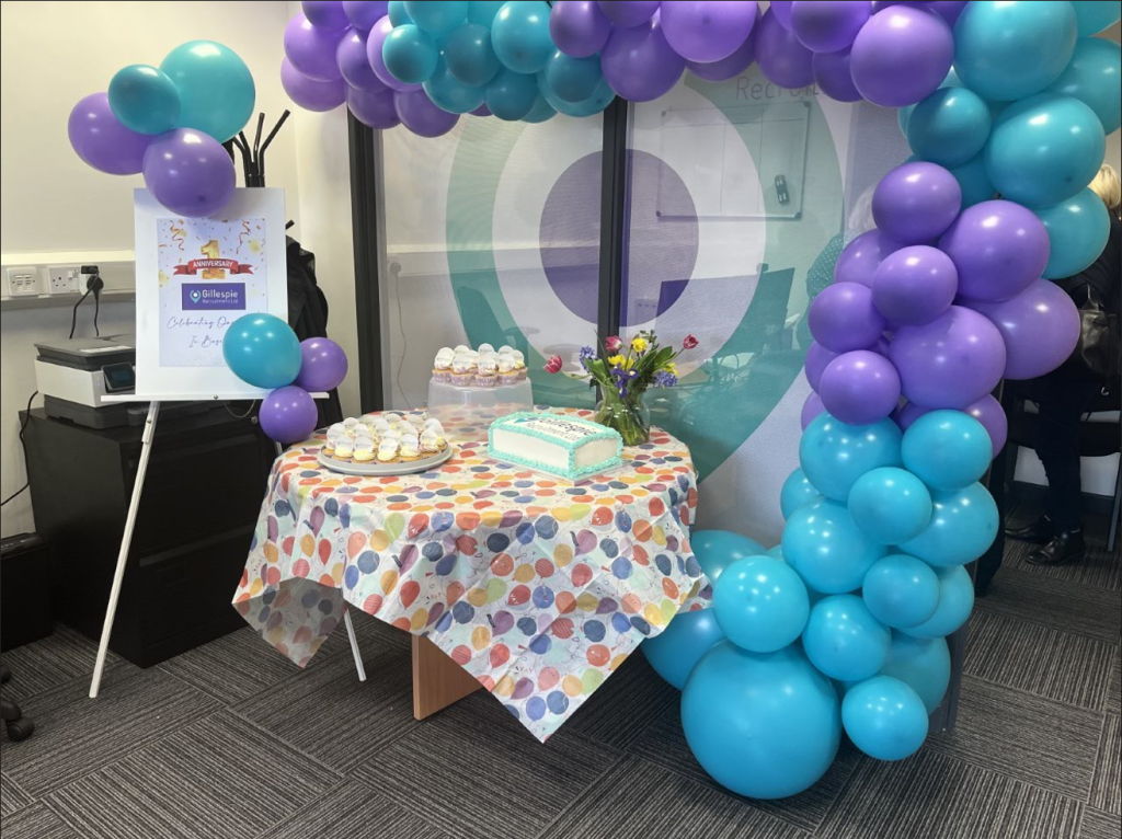 A picture in our office. The room is decorated with purple and blue balloons. The table is decorated with a spotty table cloth and there is a birthday cake and cupcakes on the table. Our party for turning one.